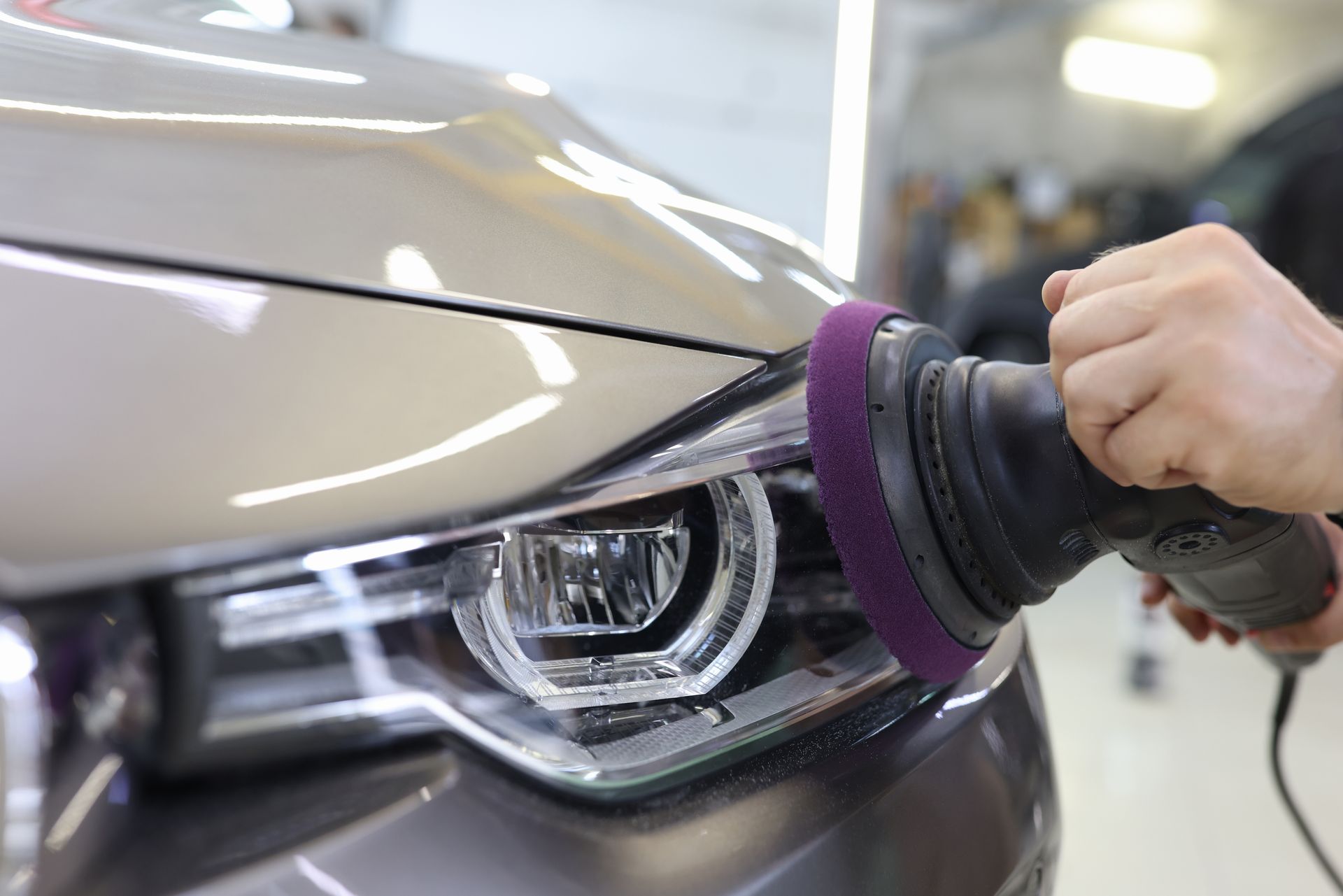 Person using a polishing tool on a car headlight, in a brightly lit garage.