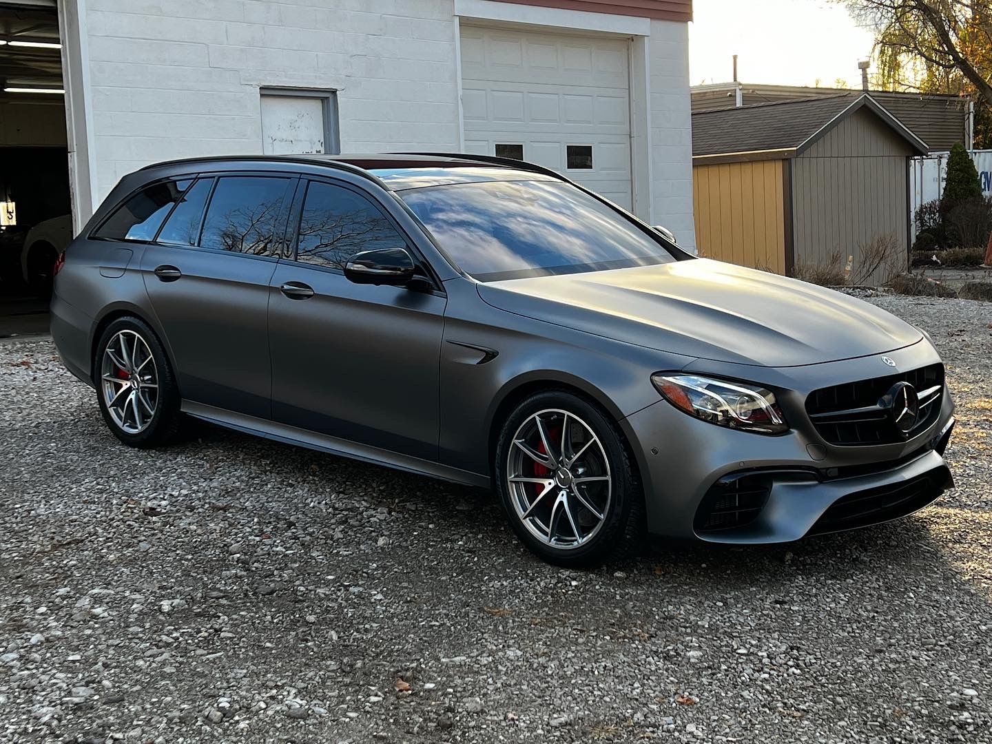 Gray Mercedes-Benz AMG E63 S wagon parked on gravel, near a building.