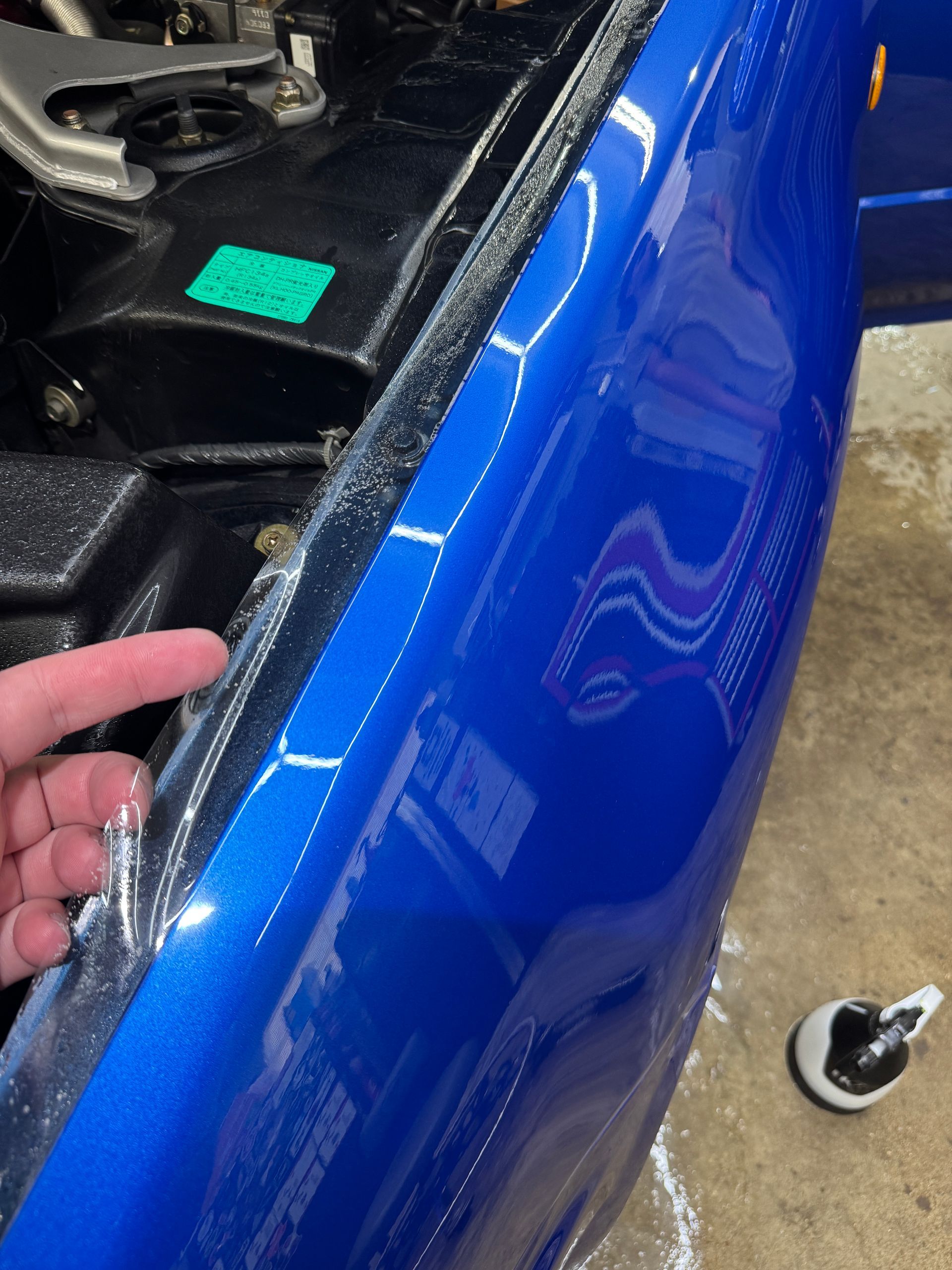 Person's finger pointing at clear film edge on blue car fender next to engine compartment.