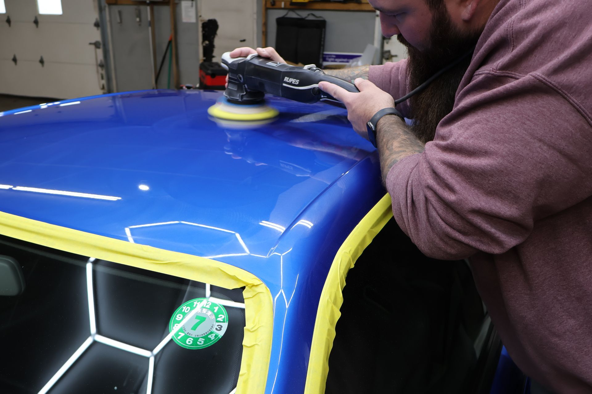 Man polishing a blue car roof with an electric buffer in a garage.