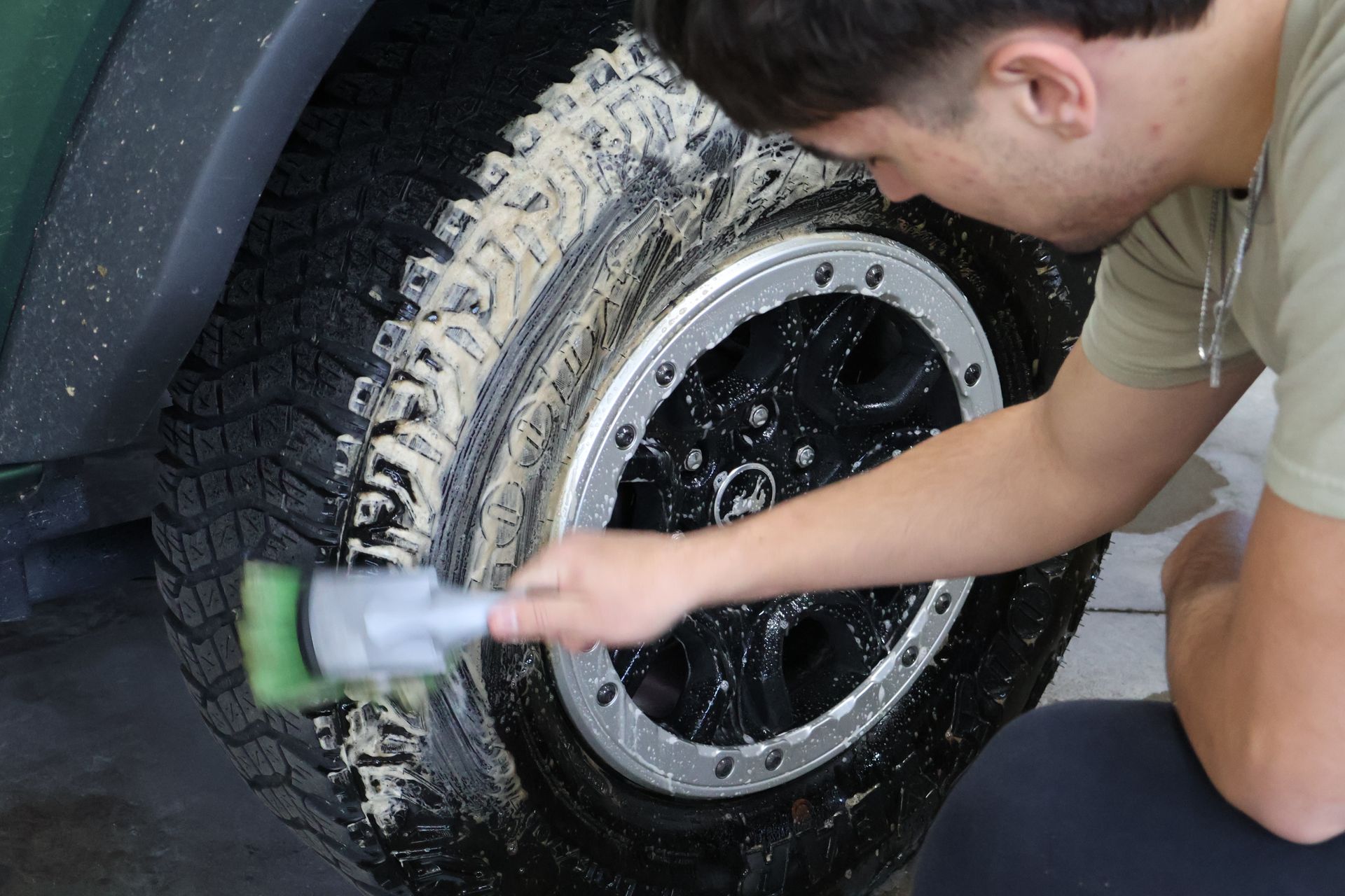 Man scrubbing a soapy car tire with a brush; wheel is black with silver rim.