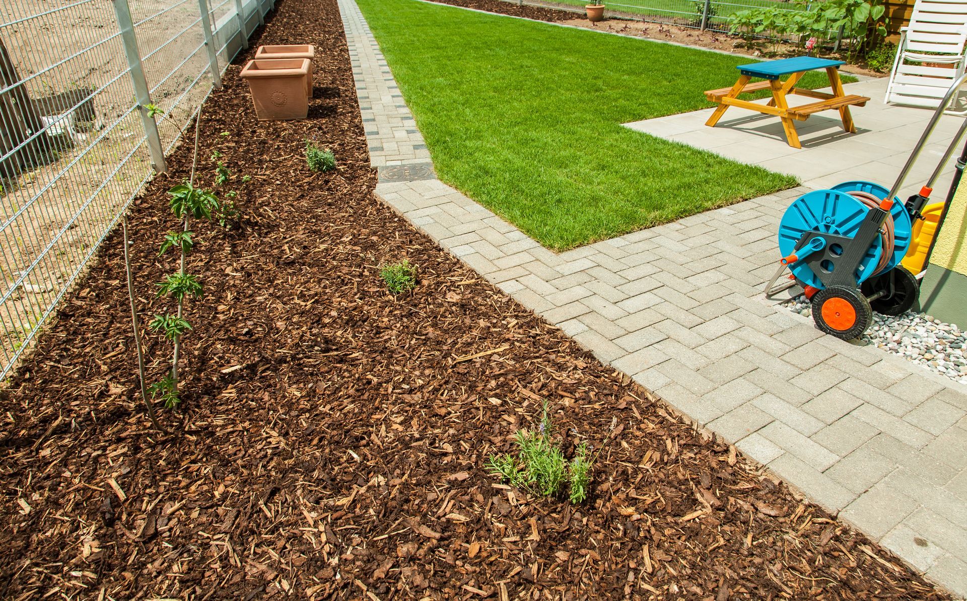 Backyard garden bed with mulch, pavers, green lawn, and a wheelbarrow near a picnic table