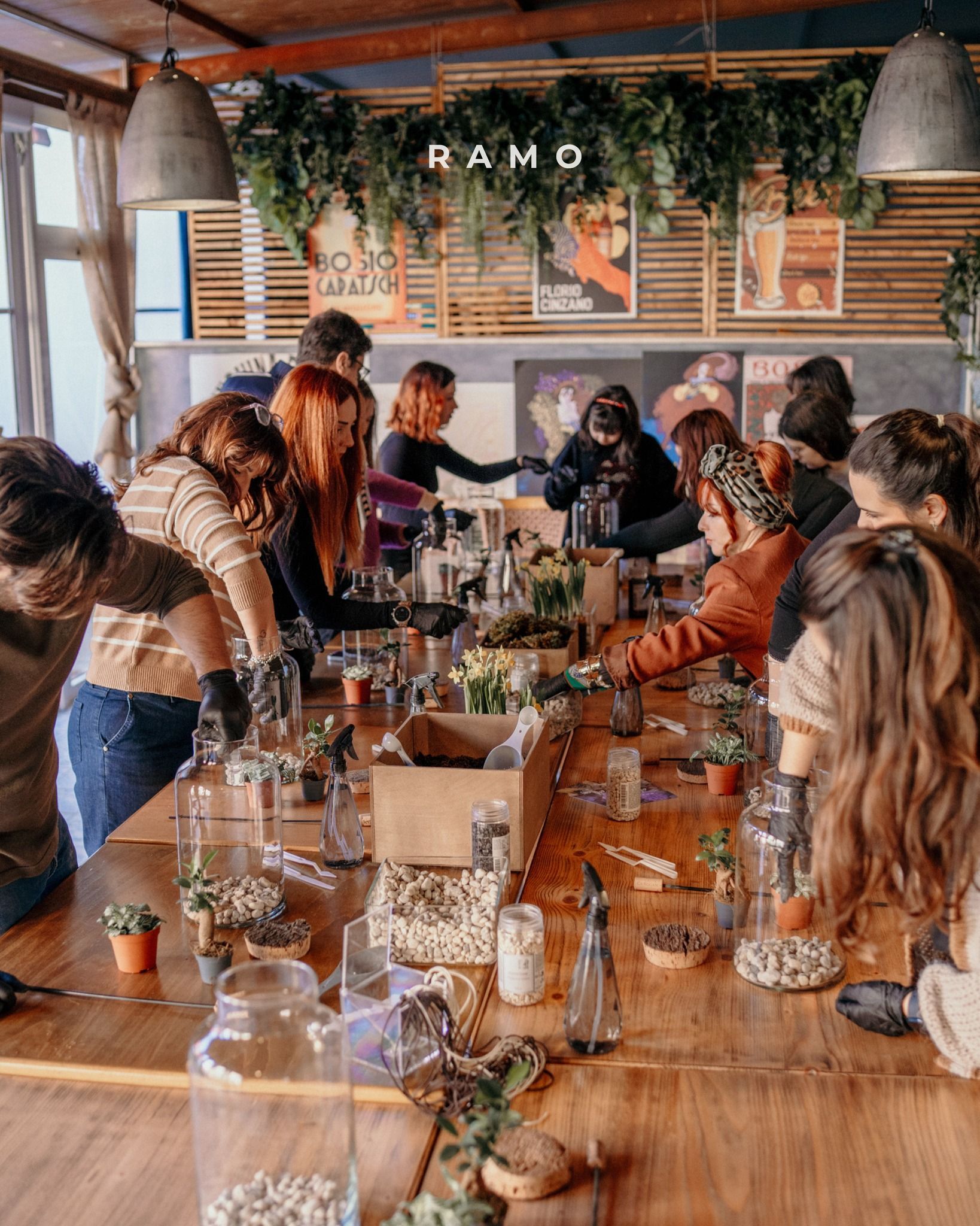 Persone che creano terrari attorno a un lungo tavolo di legno; piante, barattoli e attrezzi sono sparsi ovunque.