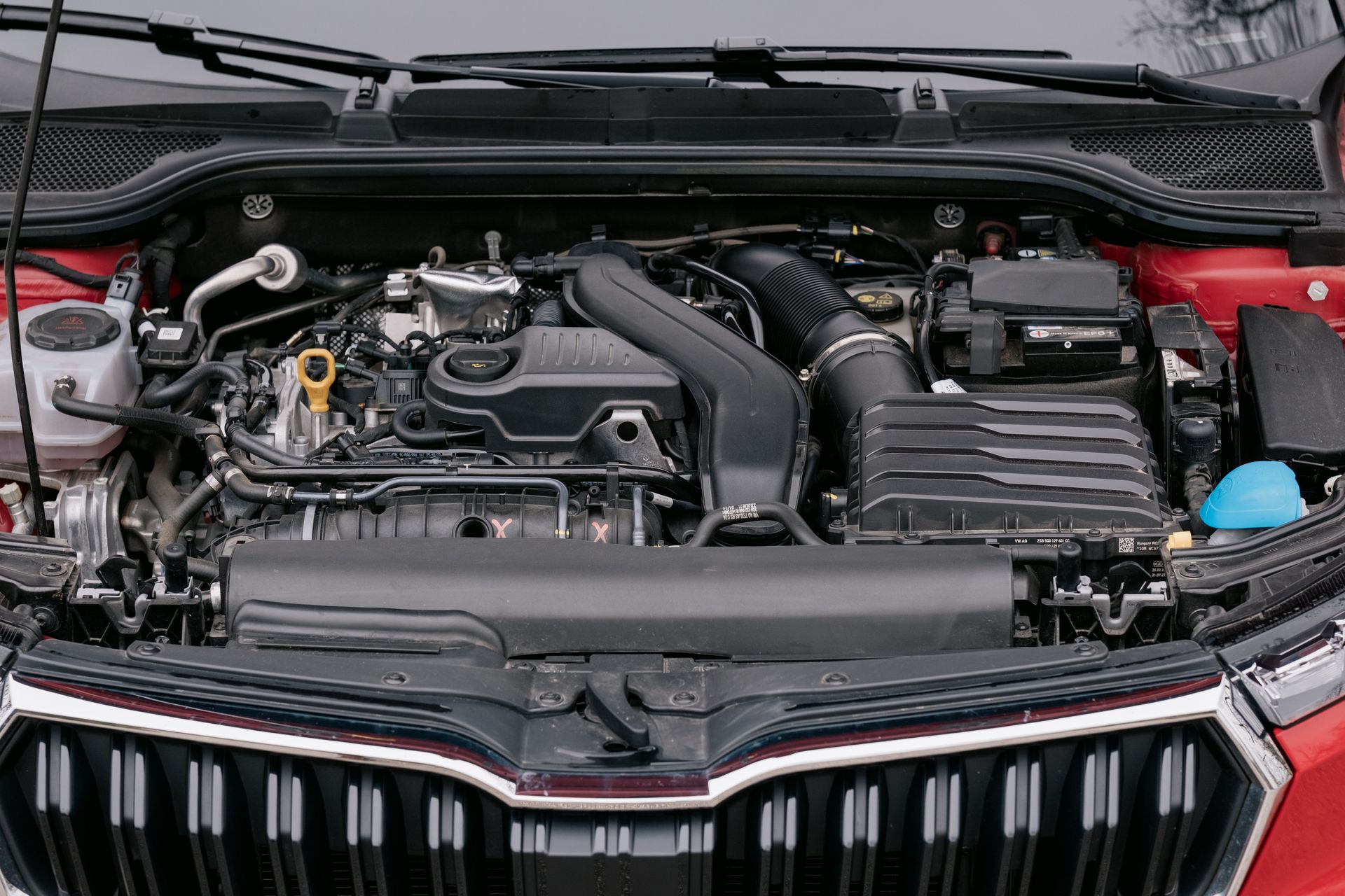 Car engine compartment, red car, components visible: air intake, hoses, battery.