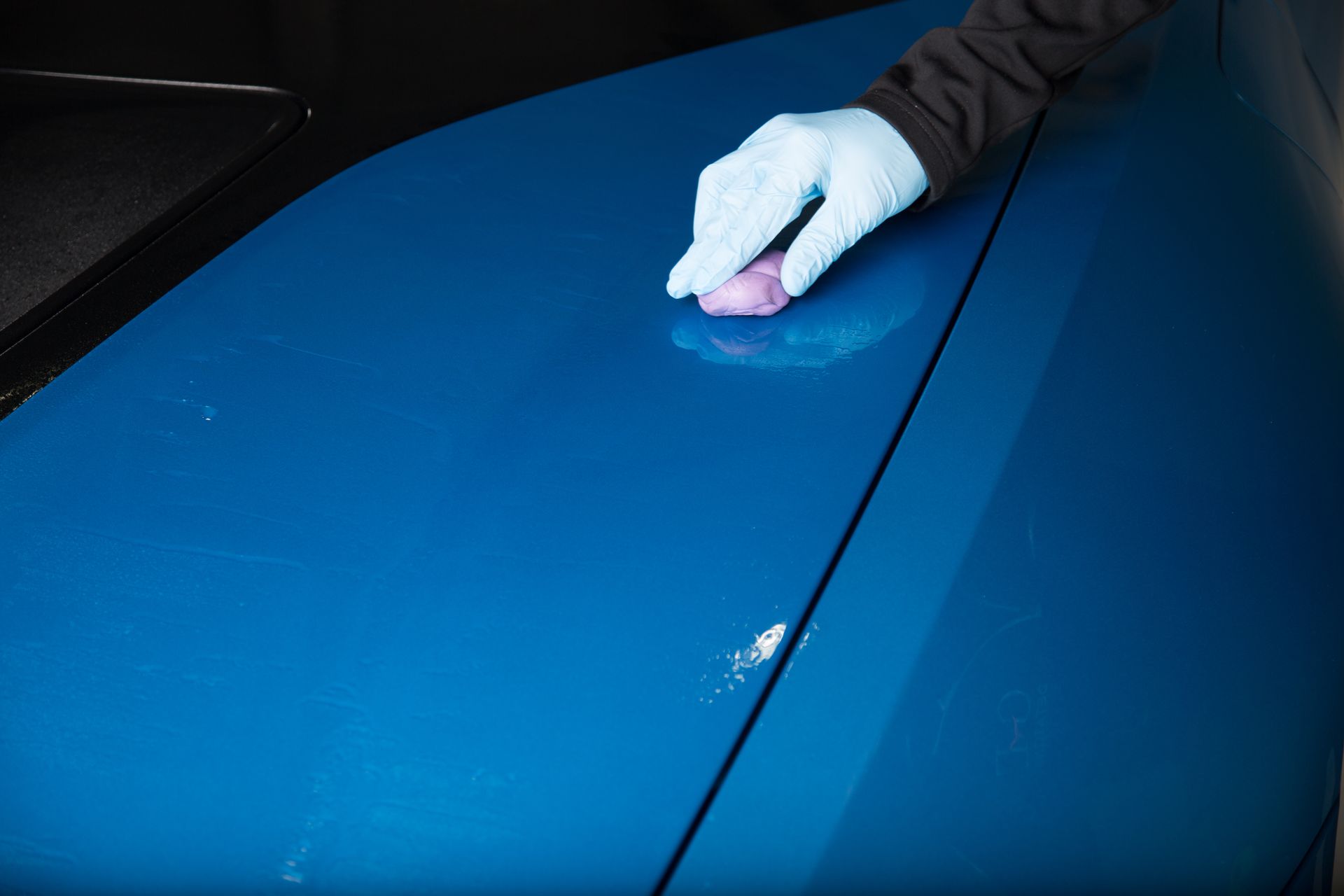 Gloved hand using purple clay bar on a blue car hood to remove contaminants.