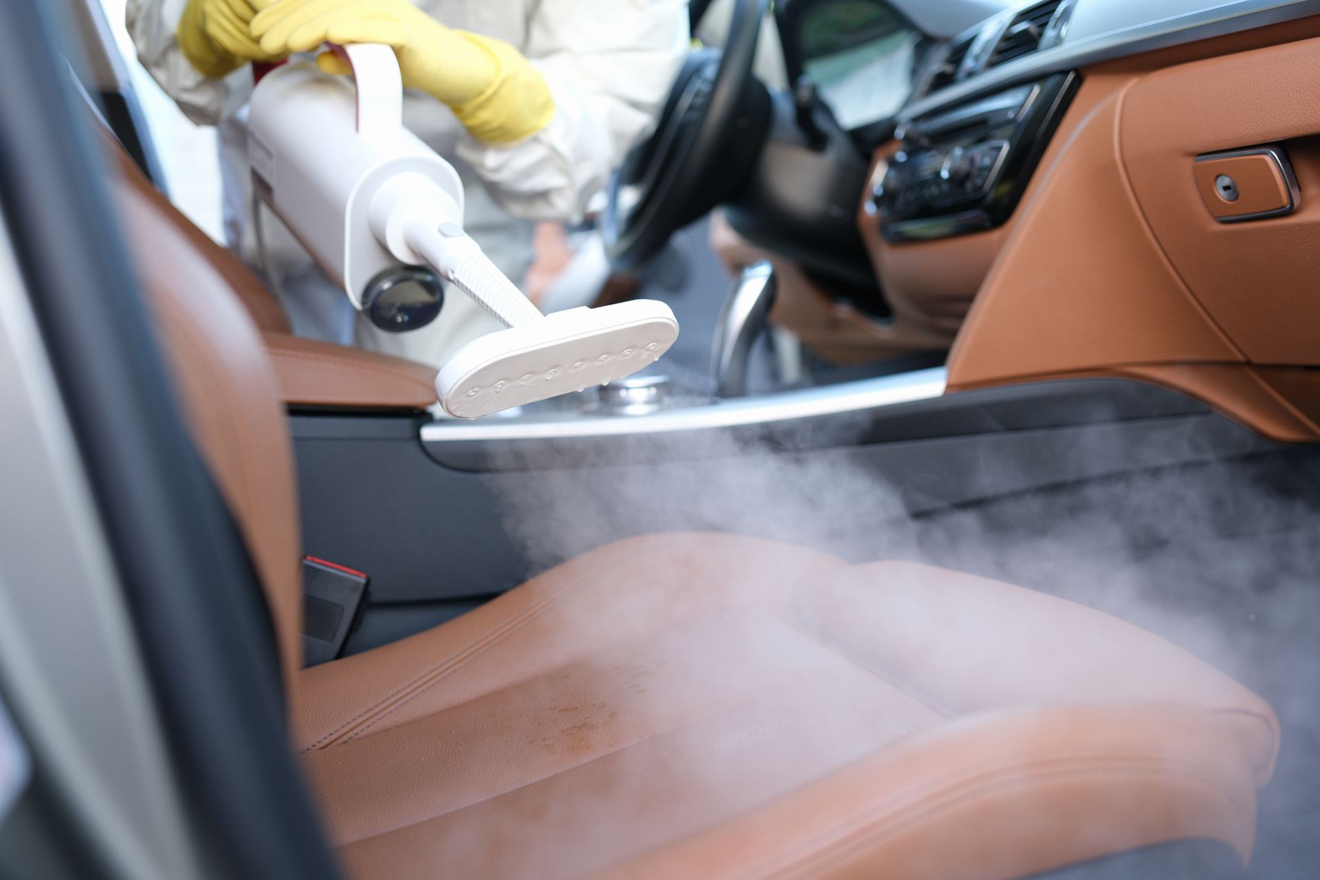 Person steaming car seat with a handheld cleaner. Brown leather interior, steam rising.