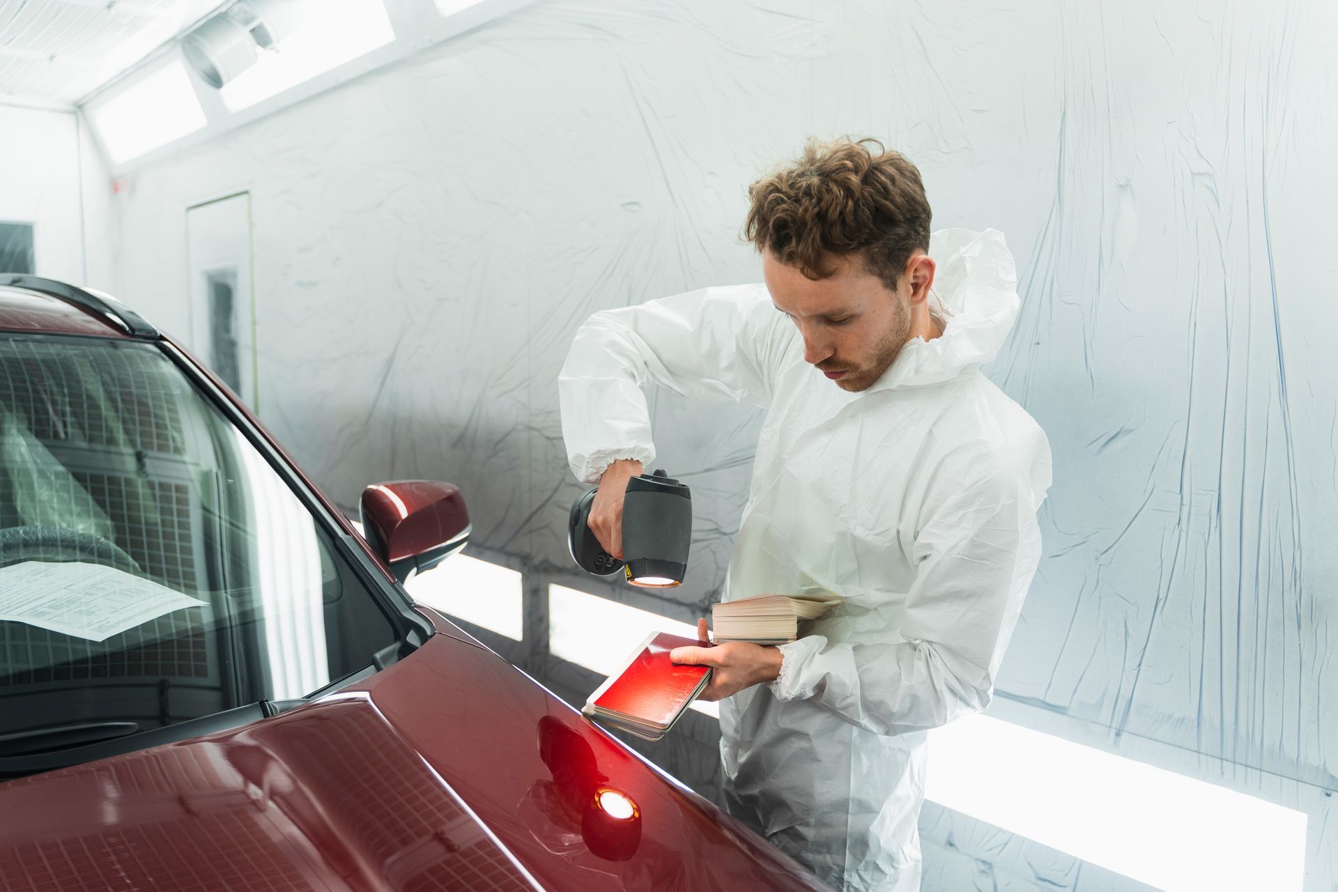 Person in protective suit using a color matching tool on a car in a paint booth.