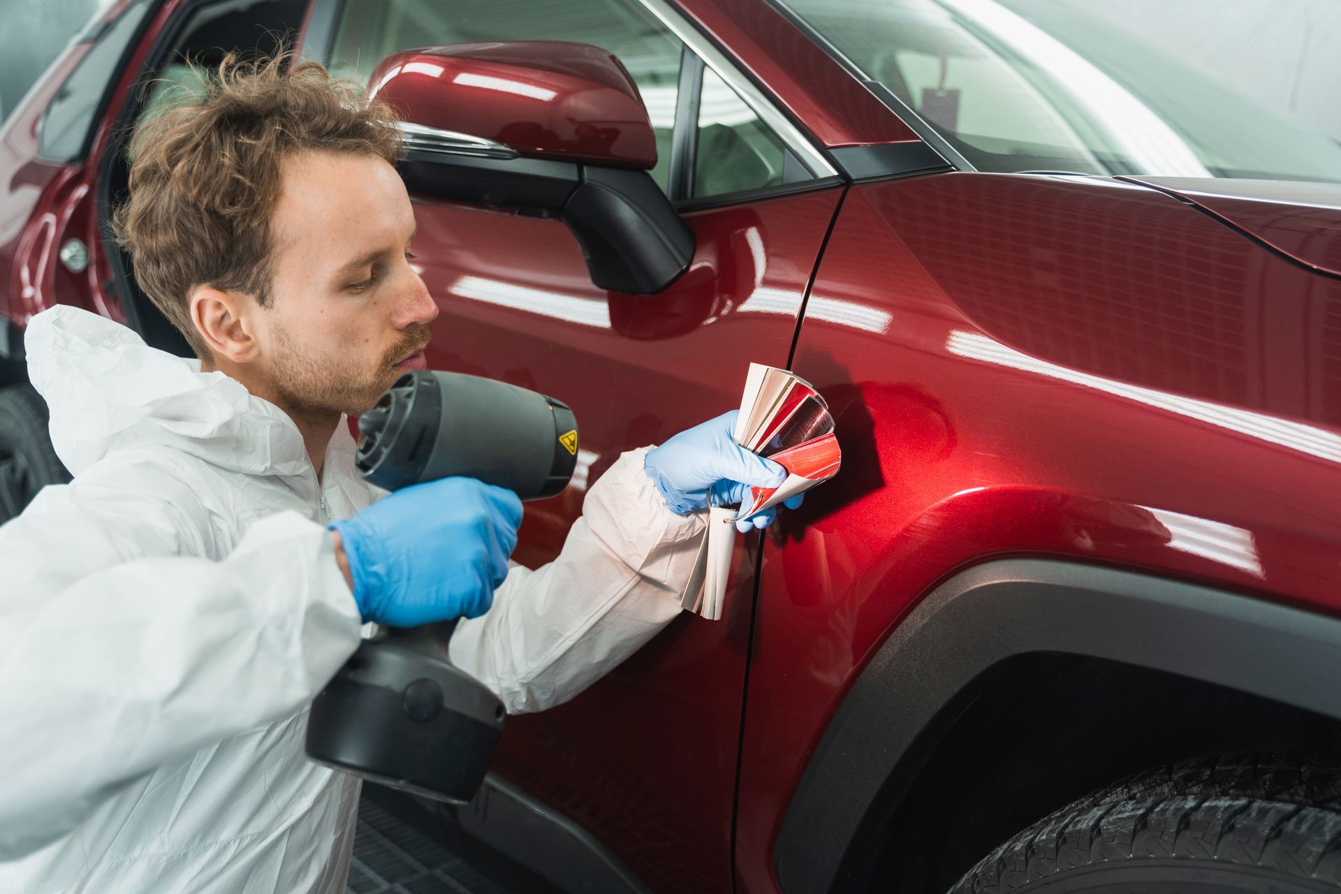 Auto body worker matching paint color with a spray gun, next to a red car.