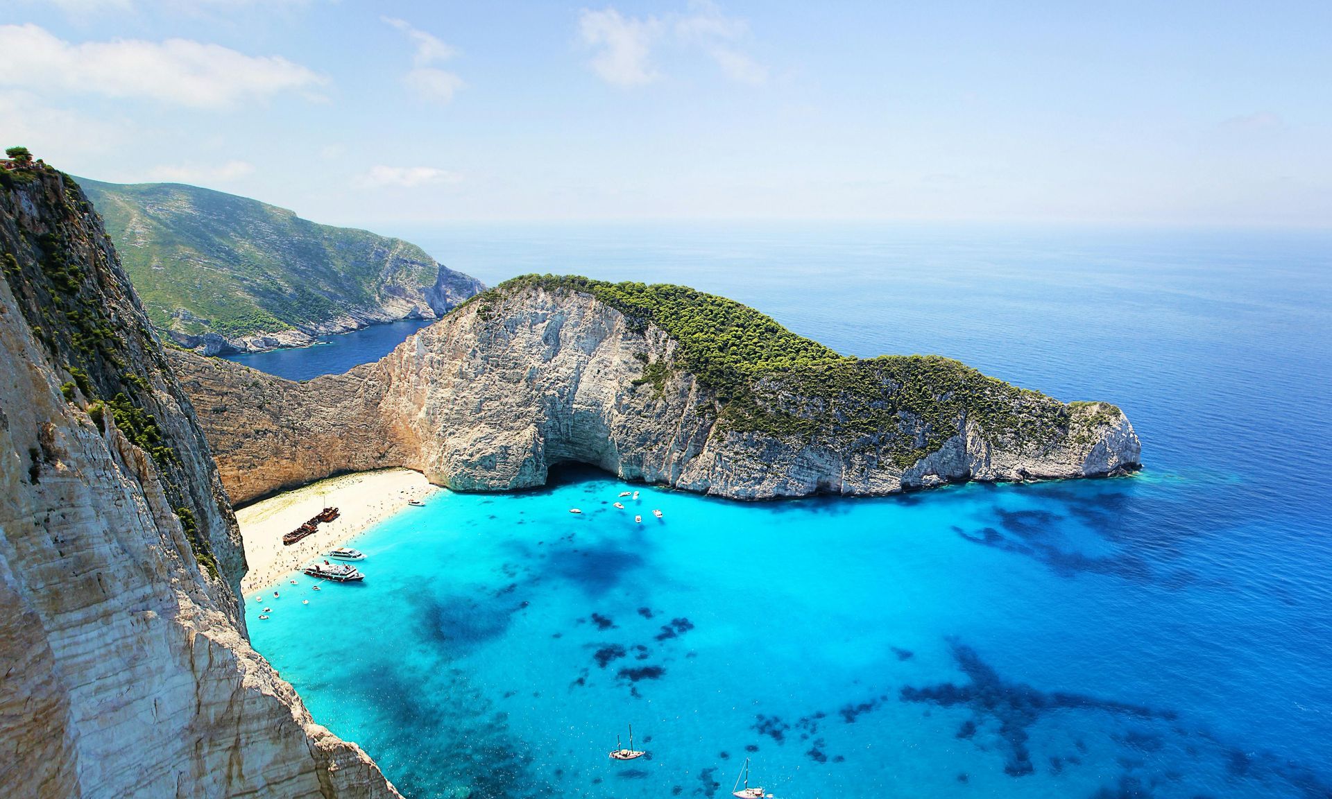 A bright turquoise cove with sandy beach, steep rocky cliffs, and clear blue sea under a sunny sky
