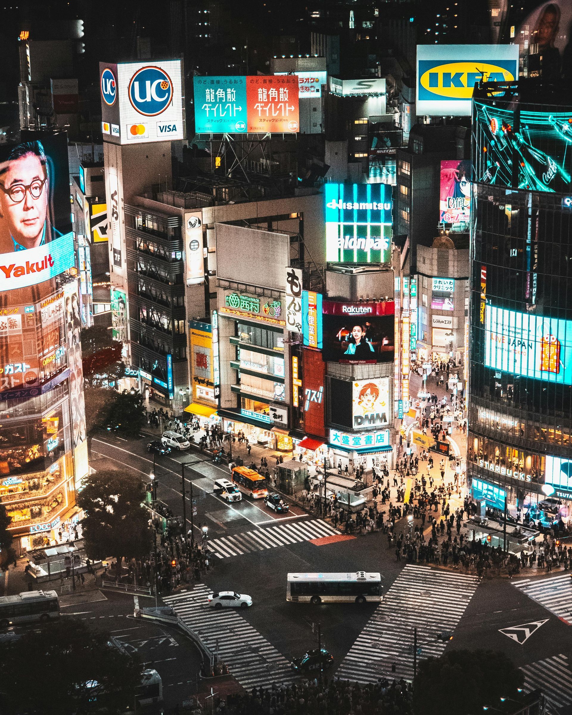 Nighttime aerial view of a busy Tokyo intersection, filled with illuminated billboards and pedestrians crossing.