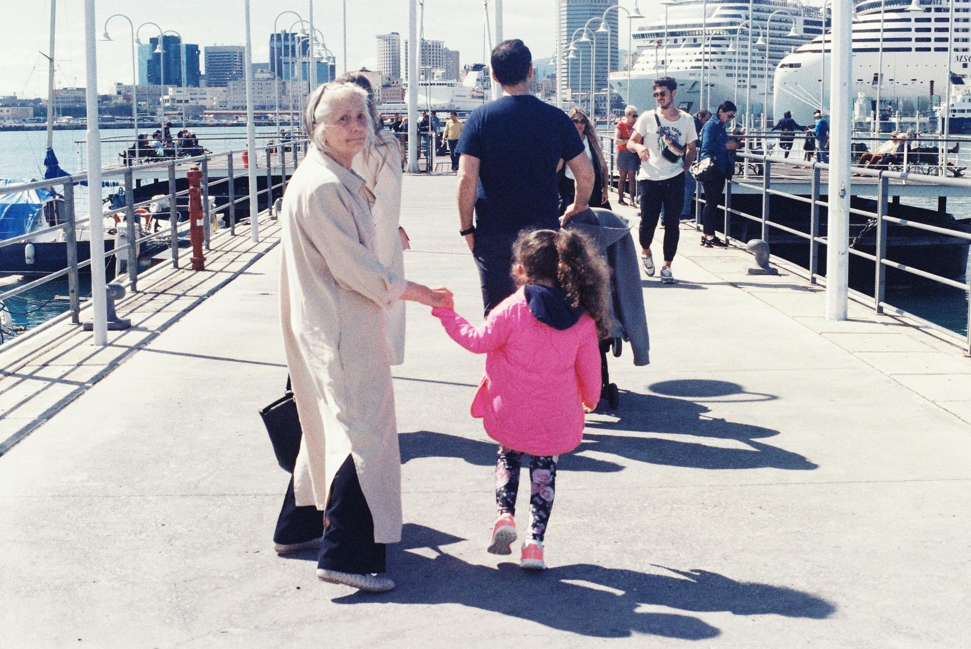 Woman holding hands with a child, walking on a pier, city and ships in background.