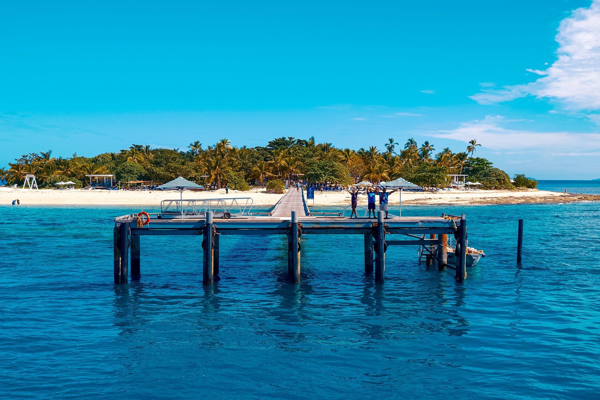 Wooden dock over bright blue water with a tropical island and palm trees in the background