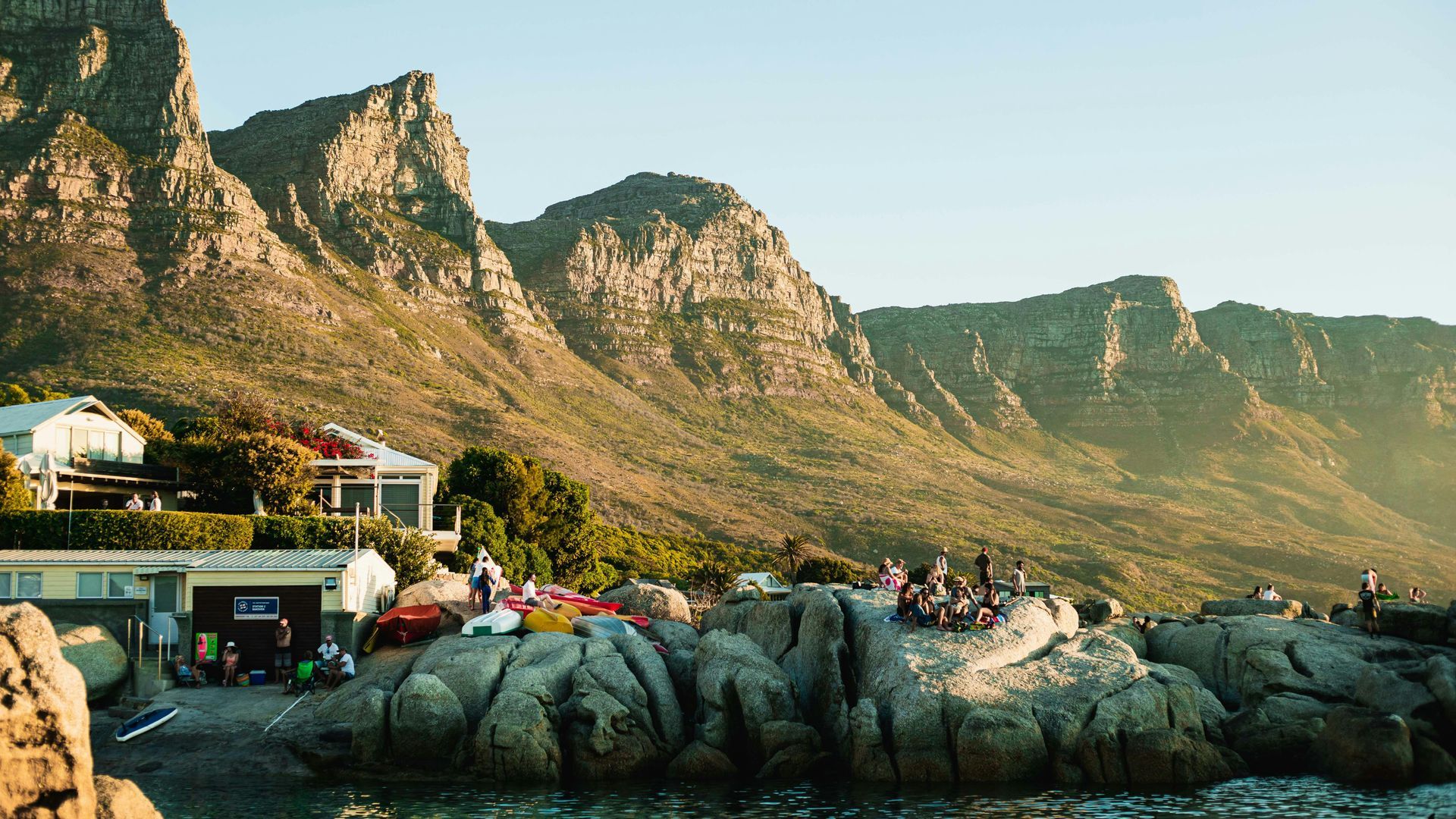 Crowded seaside rocks with a beachside café, mountain backdrop, and people watching the sunset.