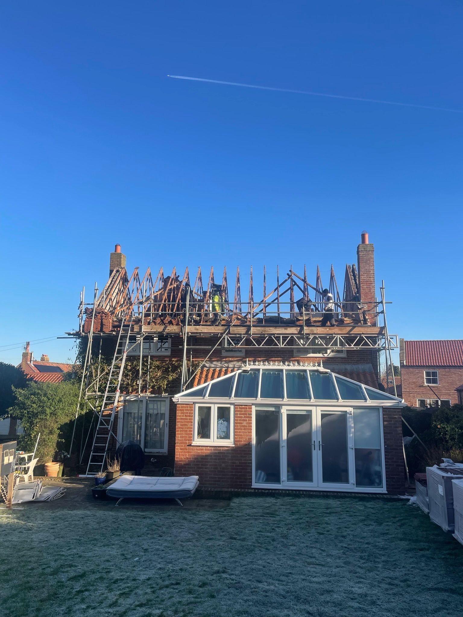 A residential house under construction, with scaffolding around the roof level, seen from a backyard on a sunny day.