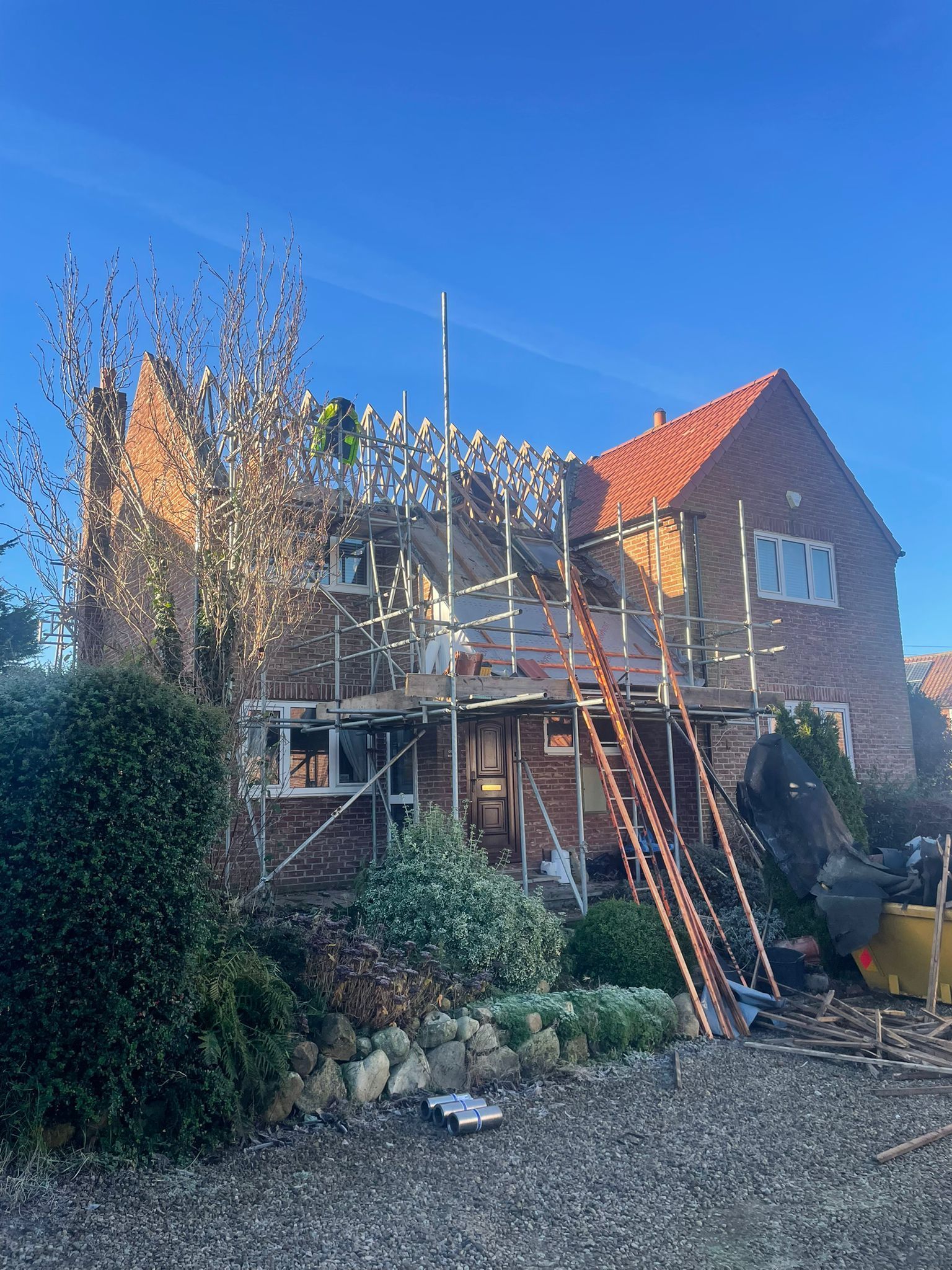 A brick house under construction, featuring scaffolding around the exterior and a bright red tiled roof under a blue sky.