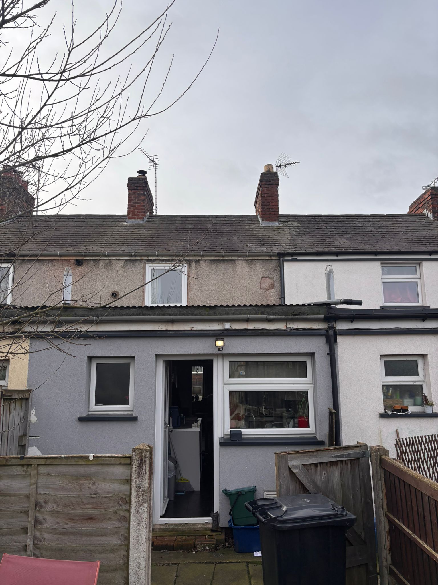 Exterior view of a terrace house back extension with grey render, a white door frame, and a slate roof under a cloudy sky.