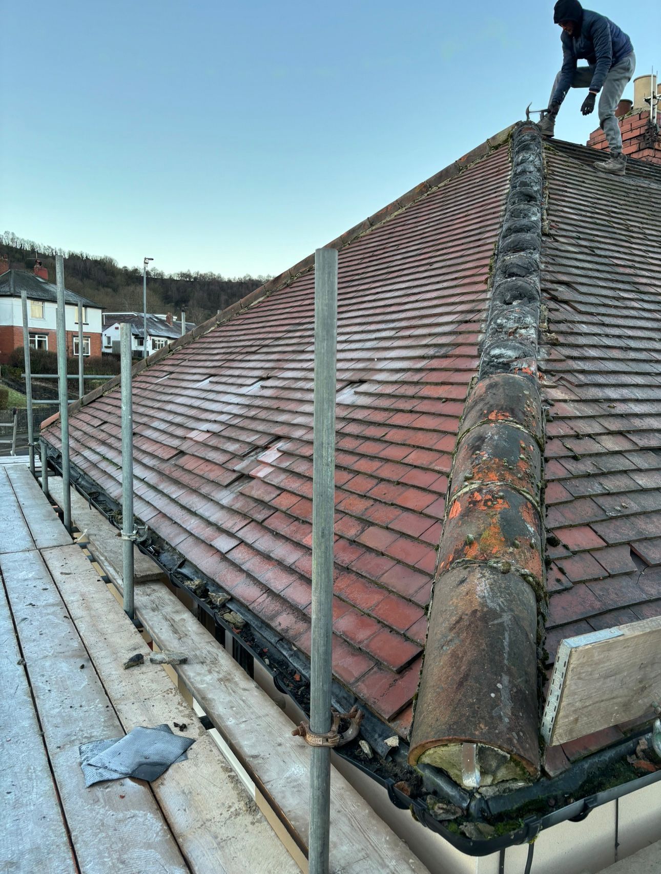 A person works on a tiled hip roof under construction, with scaffolding installed along the side of the building.