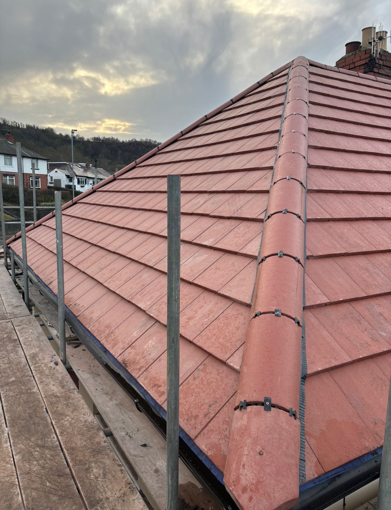A close-up view of a newly tiled red pitched roof with hip tiles, framed by scaffolding on a cloudy day.