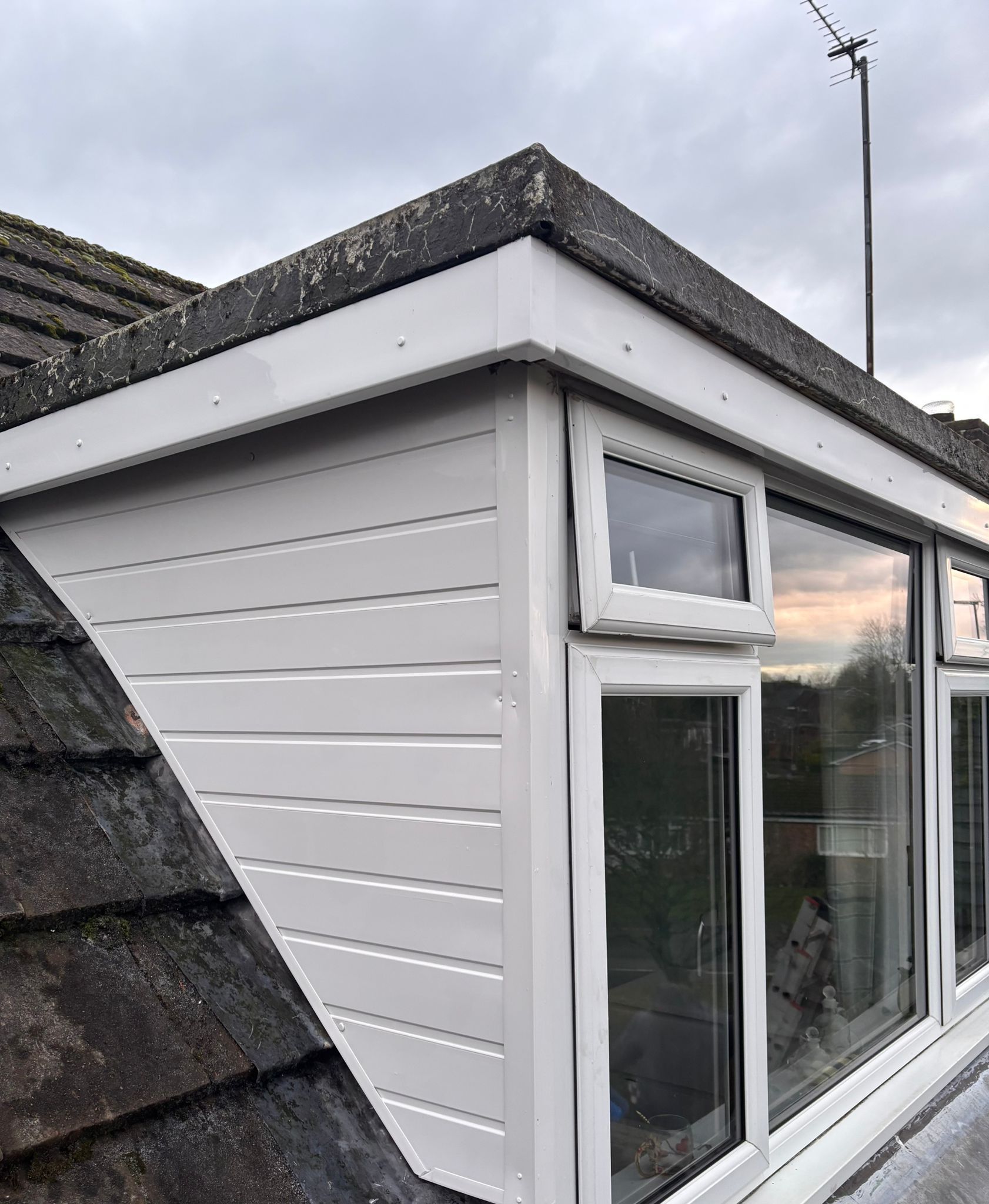A side view of a white, horizontal-sided dormer window with a grey felt roof, set against a dark tiled house roof.