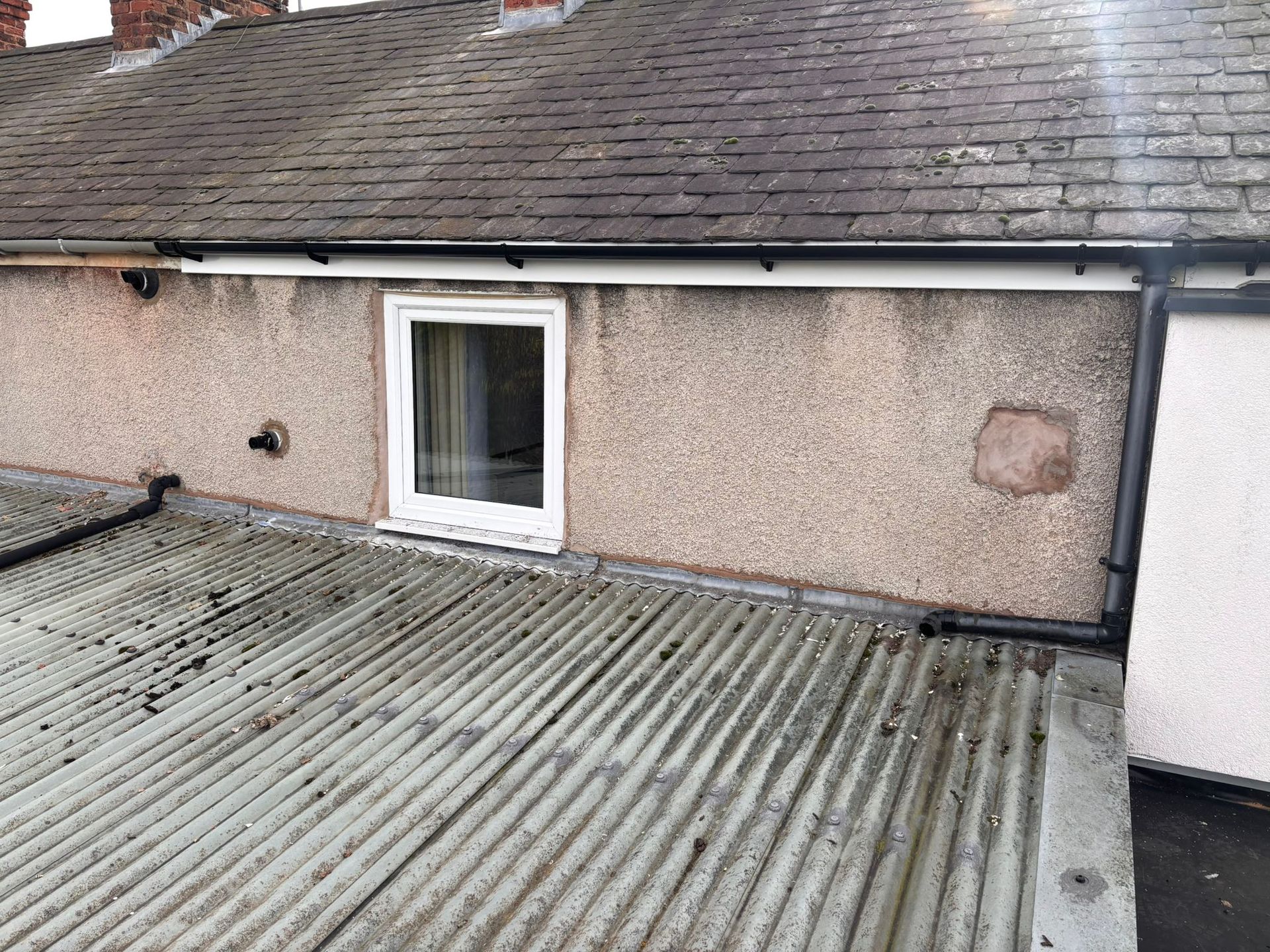 An exterior view showing a white window on a textured, beige wall above a corrugated roof with black piping and guttering.