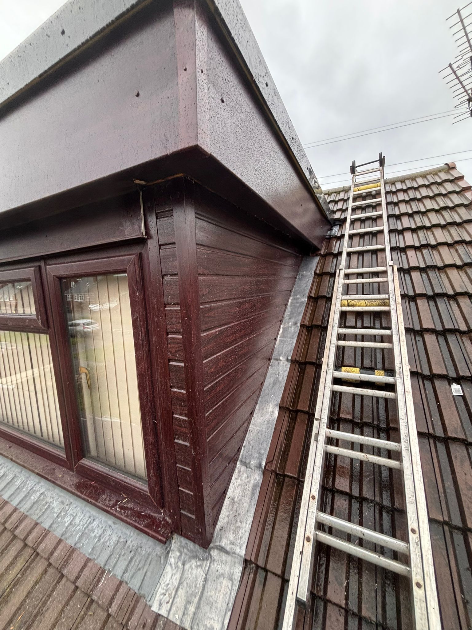 A person stands on a ladder working on the roof of a single-story brick extension attached to a two-story brick house.