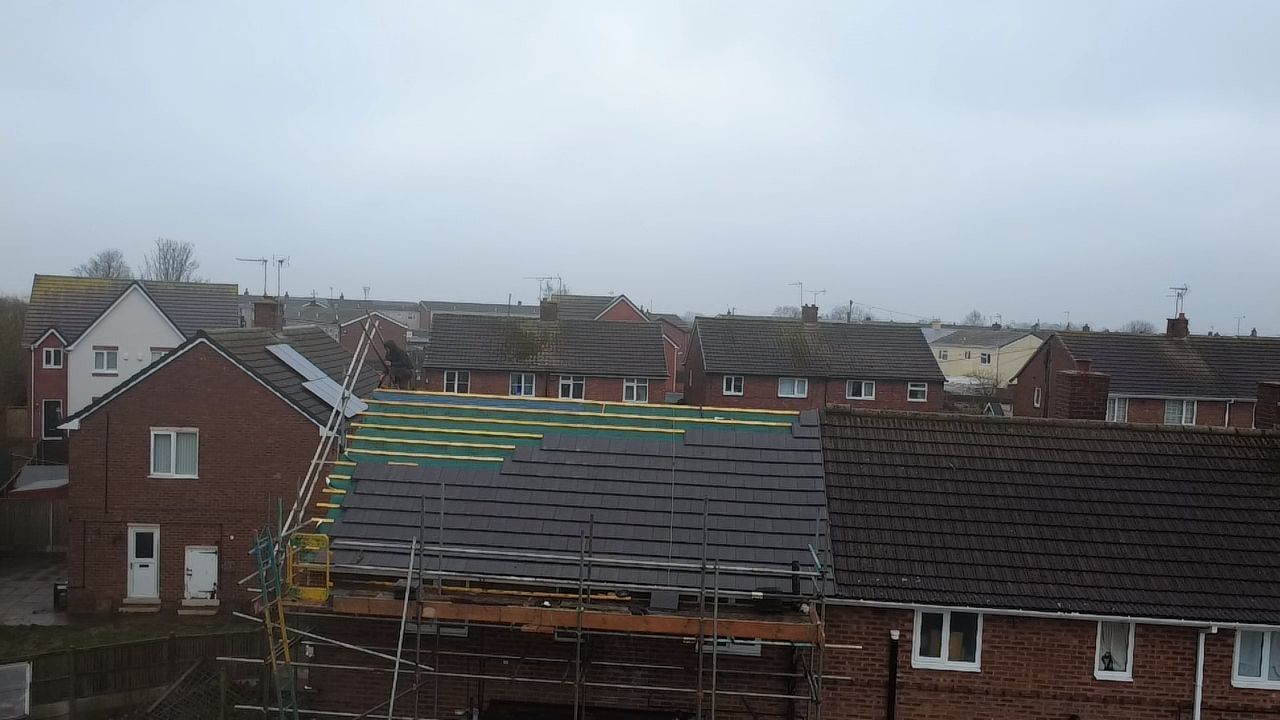 A residential street scene with a house in the foreground undergoing roof repairs, featuring exposed underlayment.