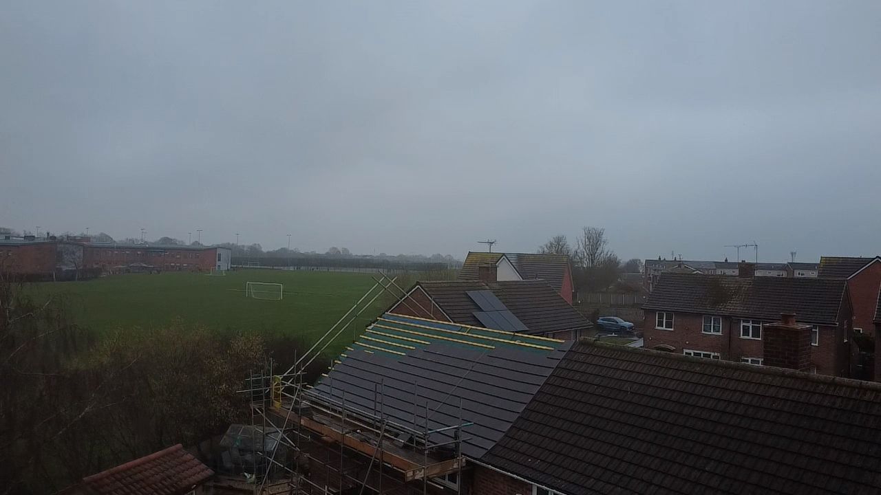 An aerial view of a building with a partially tiled roof, neighboring houses, and a green field under a cloudy sky.