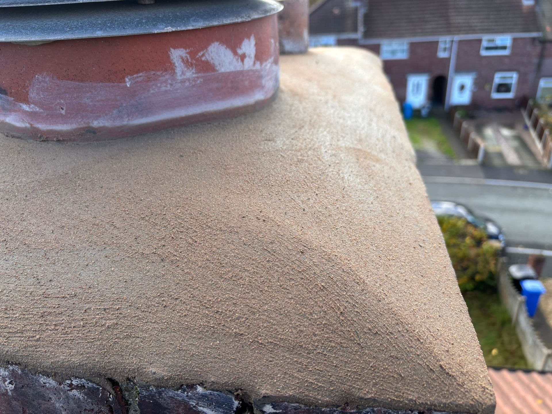 Close-up of a textured, beige concrete chimney crown with a red chimney pot, overlooking a residential street.