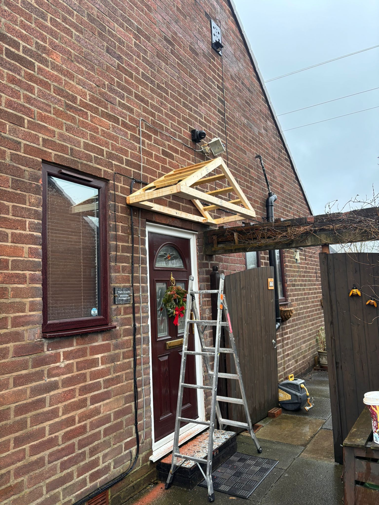 A wooden canopy frame is partially mounted above an exterior brown front door on a brick house, with a ladder in front.