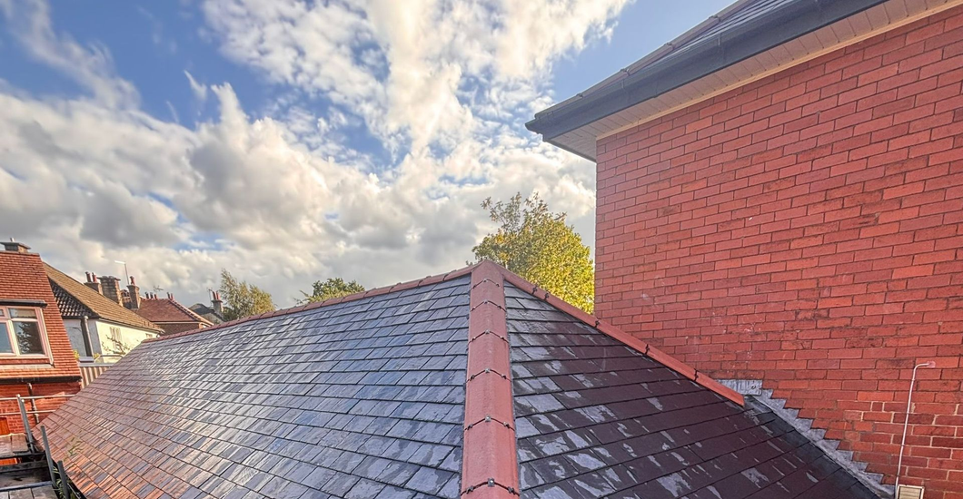 A newly laid grey slate roof with a red ridge tile, seen from scaffolding against a red brick building and cloudy sky.