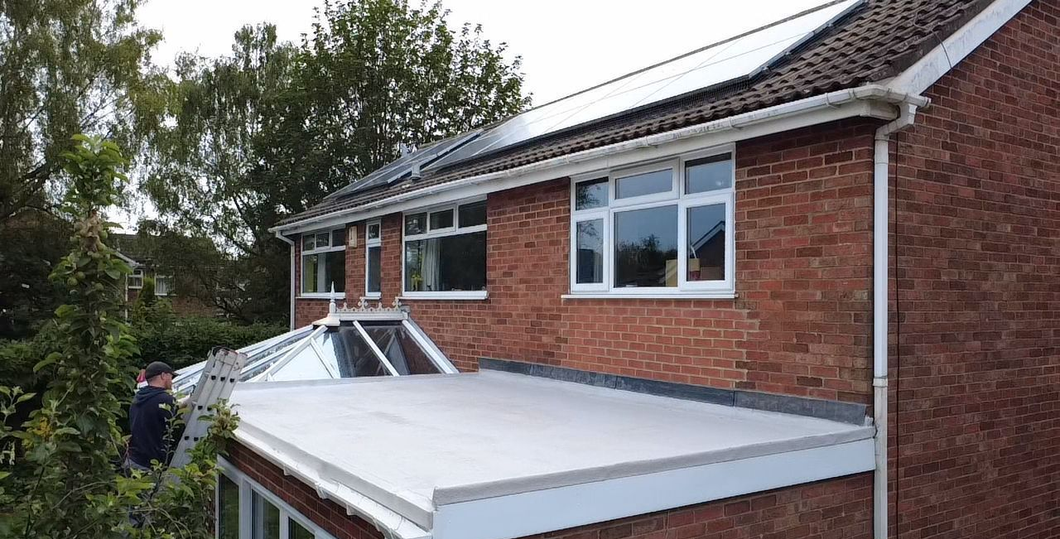 A worker stands on a ladder next to a house with a newly installed flat gray roof and a white conservatory frame.