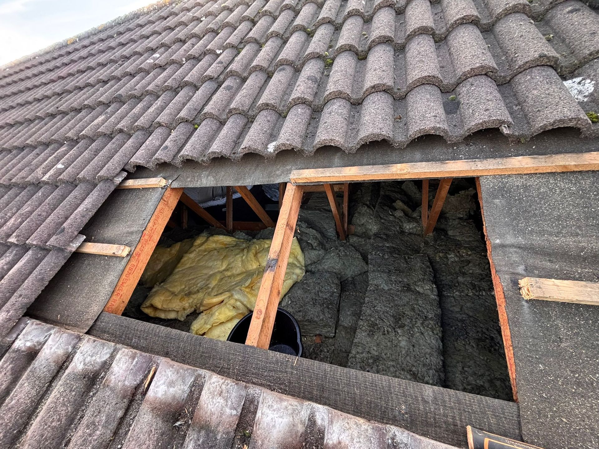 A hole in a tiled roof exposing wooden attic joists and yellow insulation material.