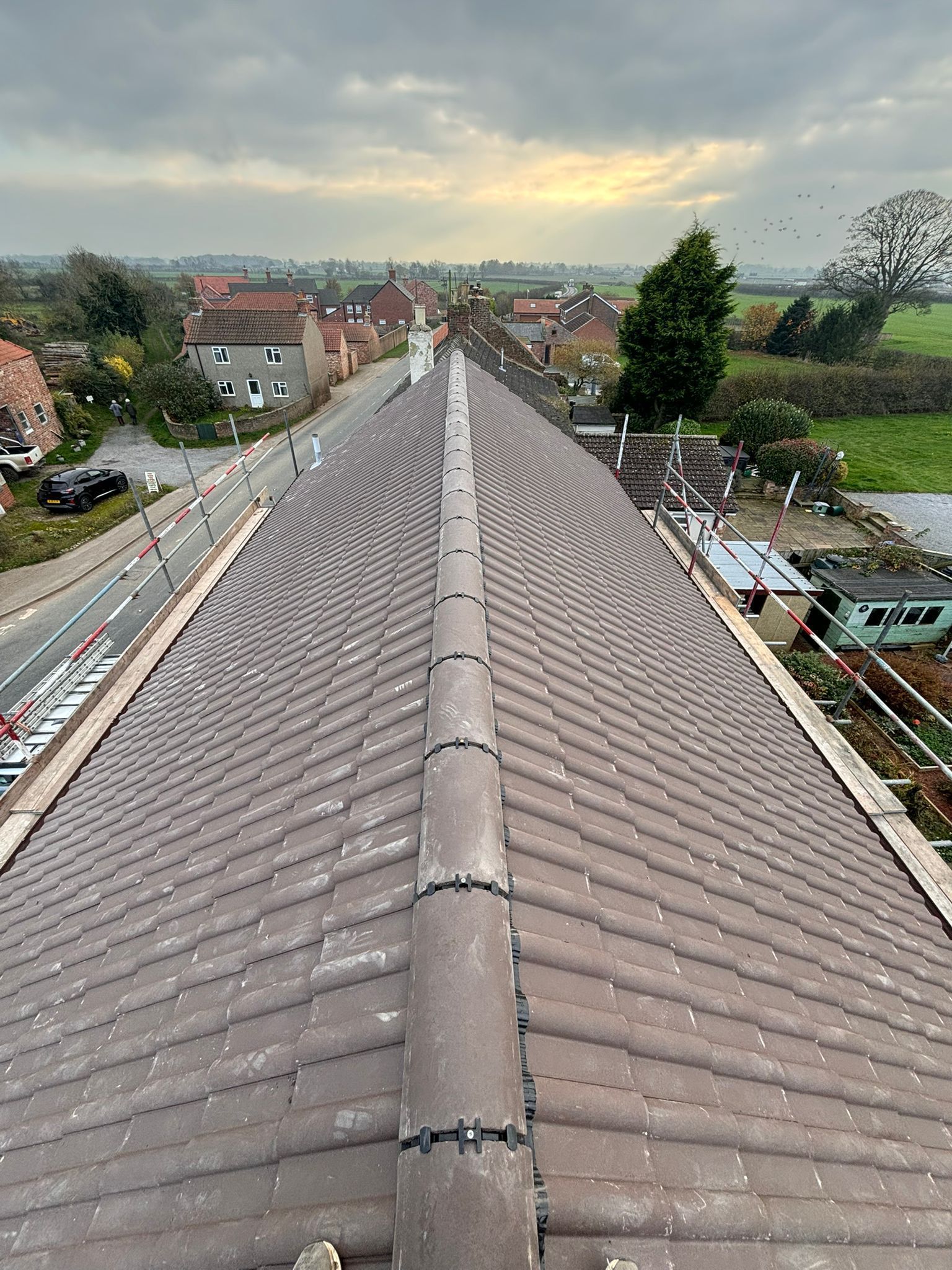 High-angle view of a brown tiled roof with a central ridge, overlooking a quiet street, houses, and fields at dusk.