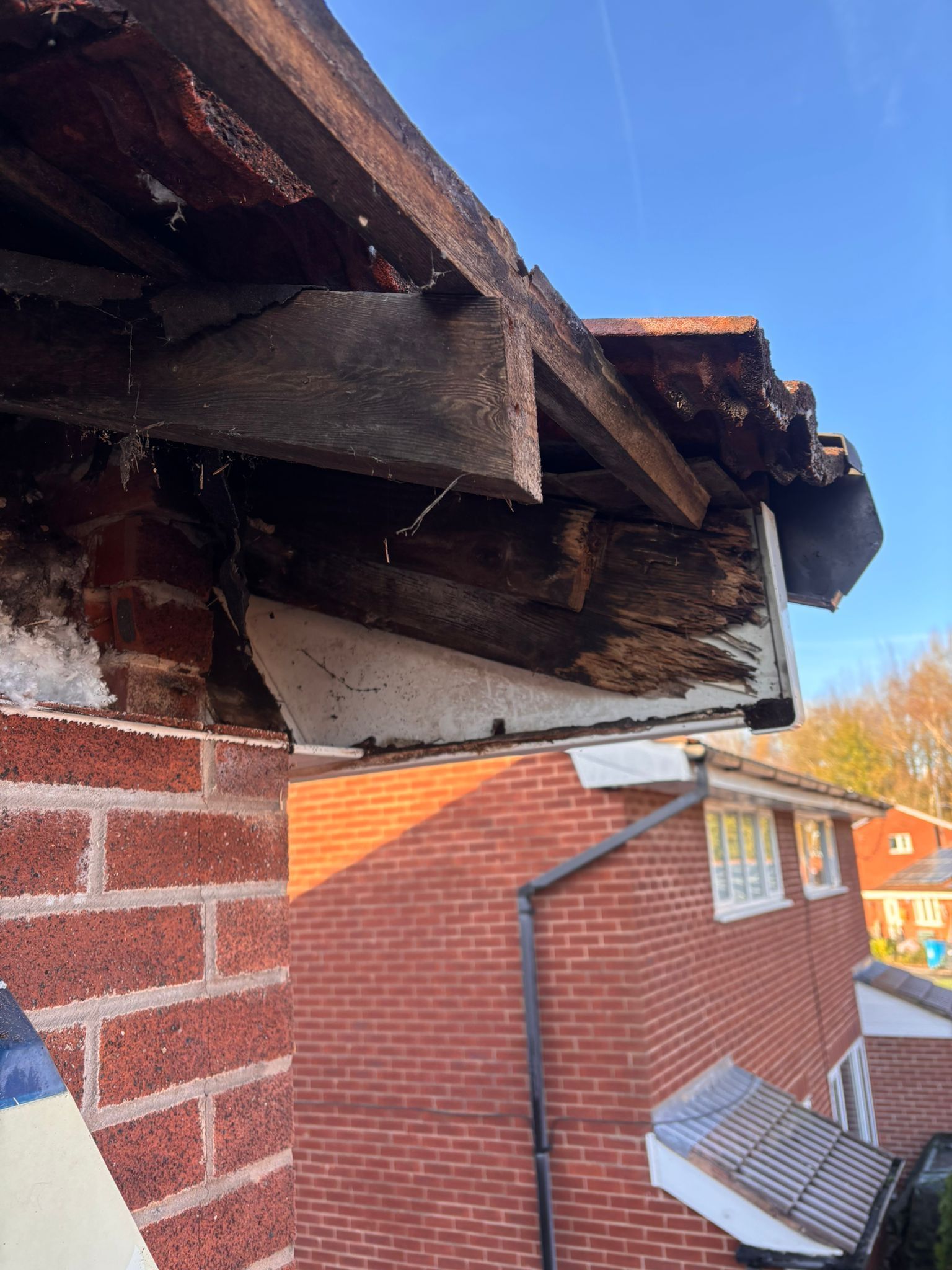 Damaged wooden roof eaves and exposed rafters on a brick house against a blue sky.