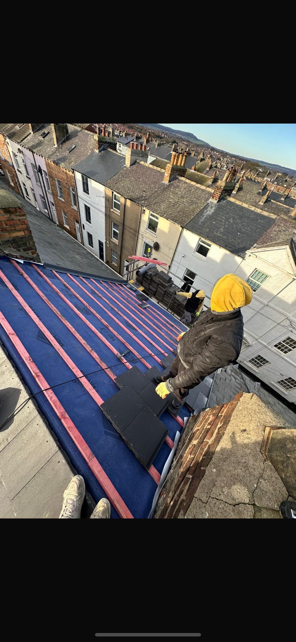 A person wearing a yellow hard hat works on a residential roof covered with blue waterproof underlayment.