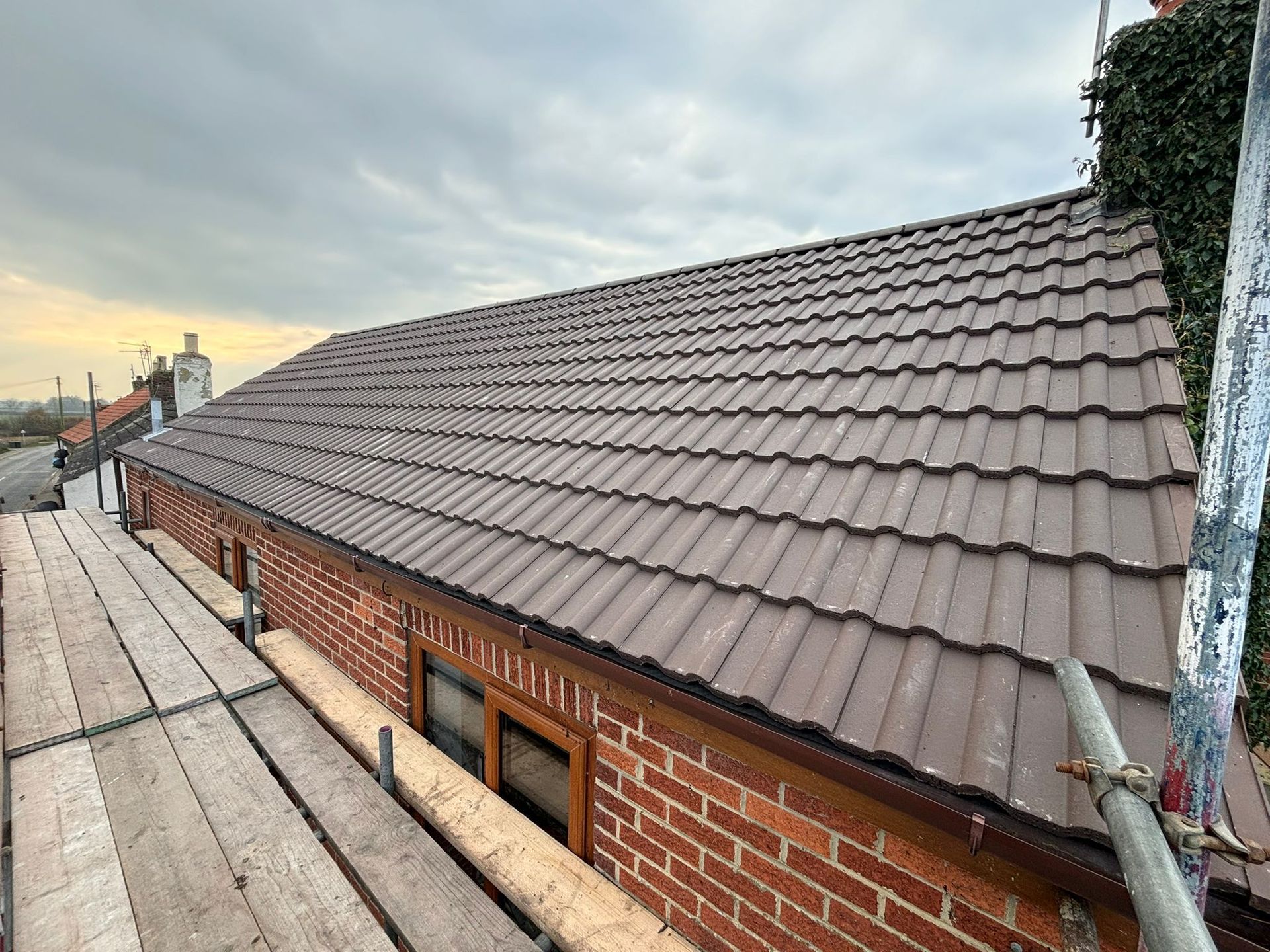 A brown tiled roof on a brick building, viewed from adjacent scaffolding under a cloudy sky.