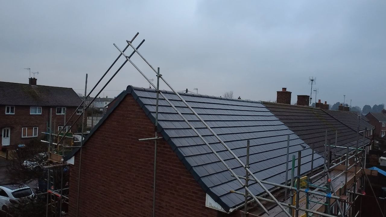 Scaffolding frames a house roof with new dark gray tiles under a cloudy sky in a residential neighborhood.