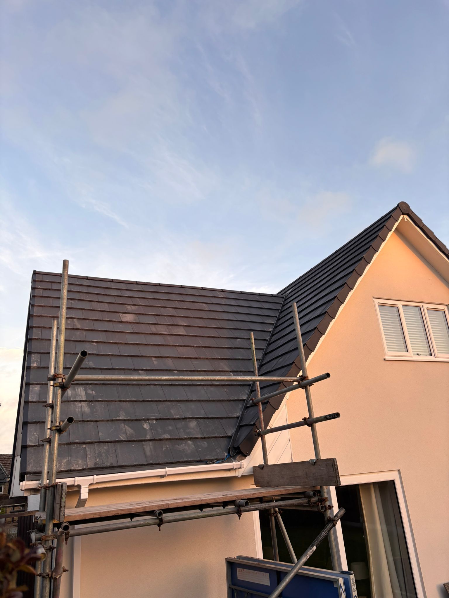 A construction scaffold stands beside a house with a dark tiled roof and a light-colored exterior wall.