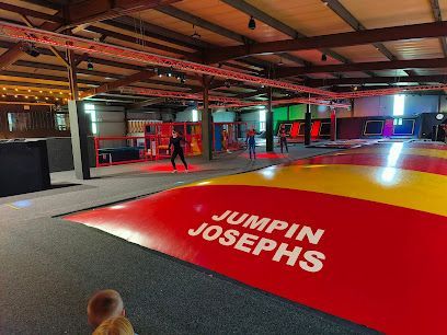 Indoor trampoline park with red and yellow mats and “JUMPIN JOSEPHS” on a ramp; person walking in background