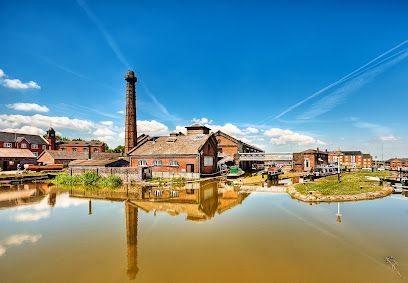 Canal-side brick buildings and a tall smokestack reflected in calm water under a blue sky