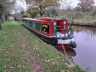 A green and red narrowboat moored beside a canal towpath with autumn trees and a bridge in the distance