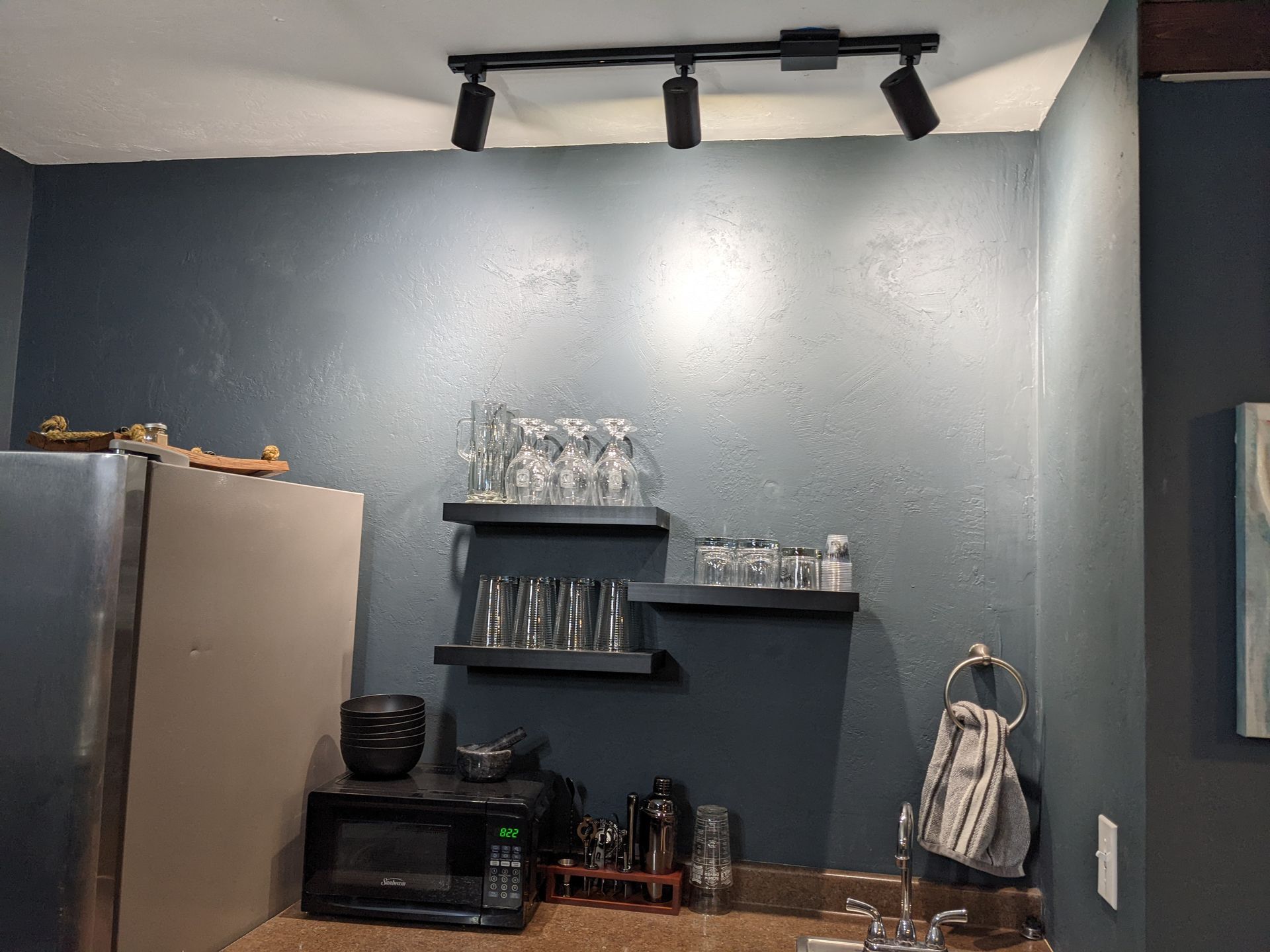 A kitchen with a refrigerator , microwave , shelves , glasses , and a sink.