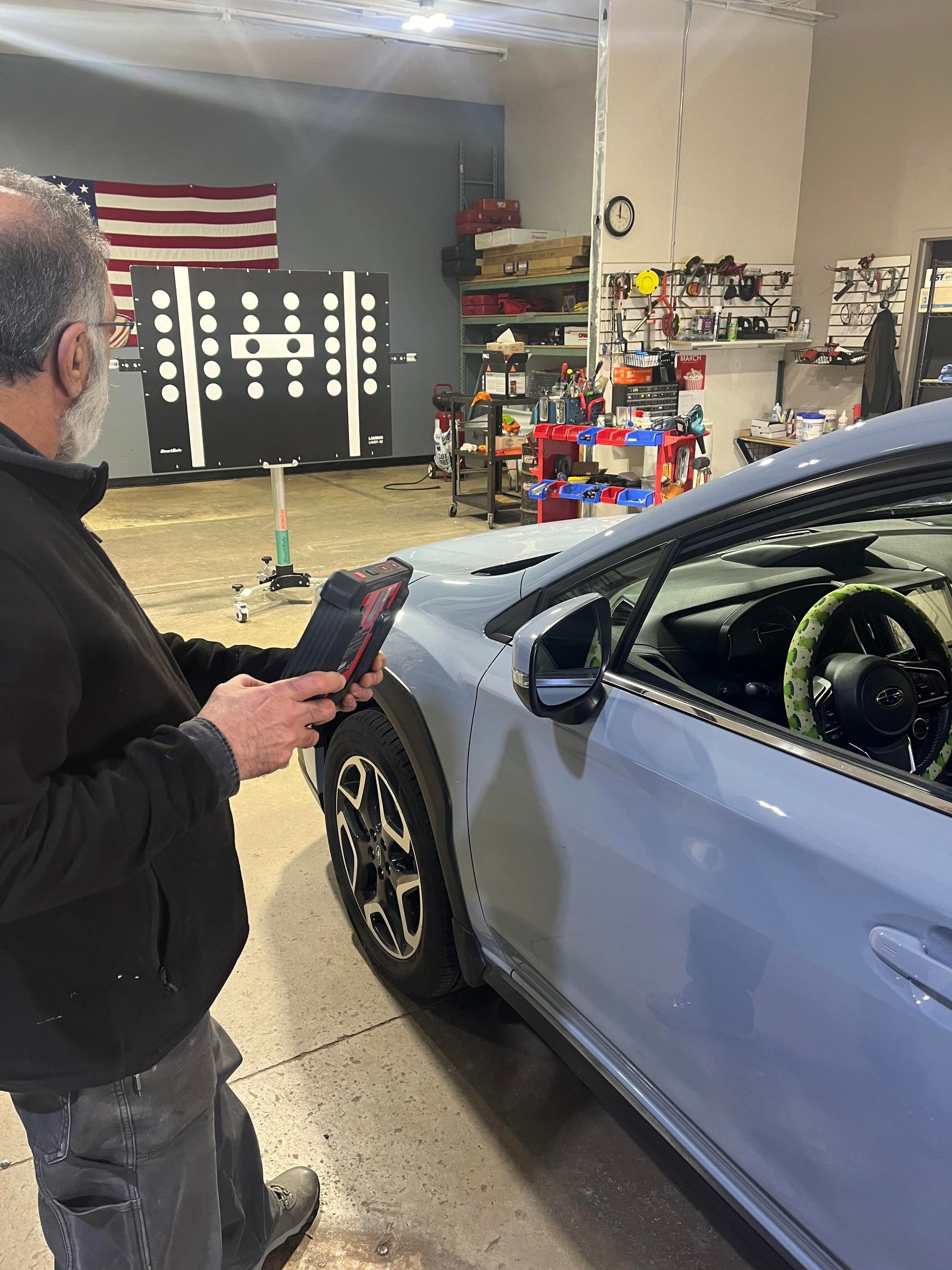 A man wearing a mask is holding a tablet in front of a car in a garage.