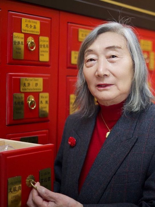 Dr Wu in a red sweater is standing in front of chinese medicine drawers