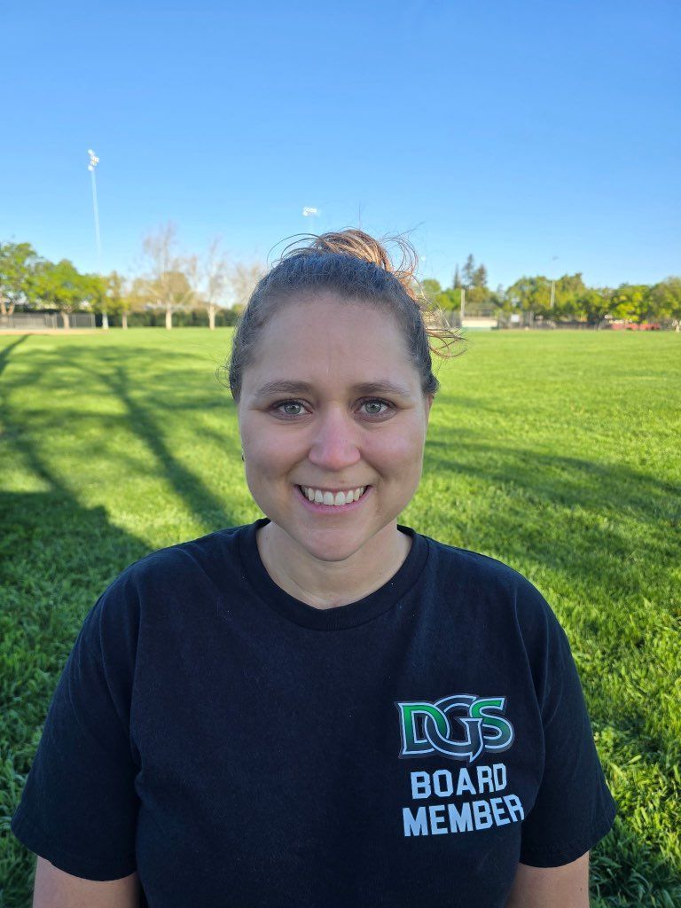 Football coach with arms crossed, standing on a football field. He wears a navy polo shirt and gray pants.