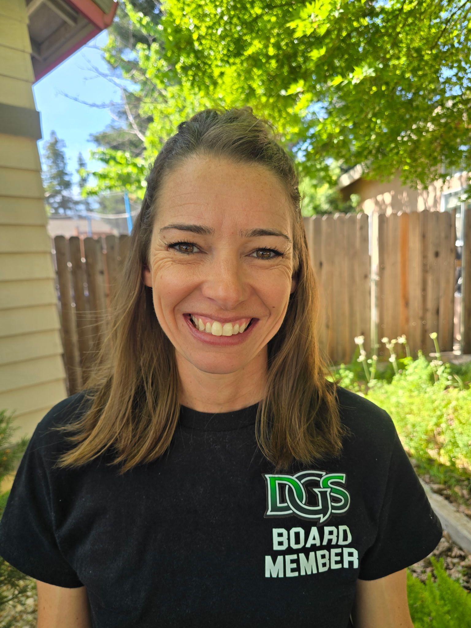 Football coach with arms crossed, looking stern on a sunny field, players in the background.