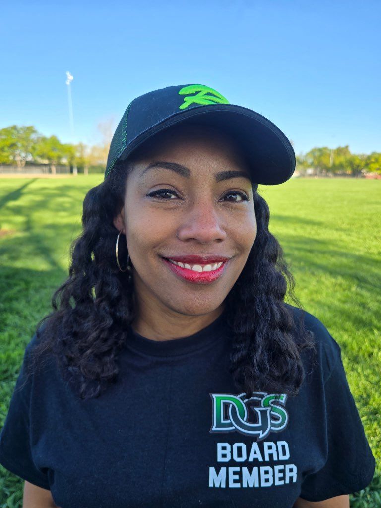 Football coach with arms crossed, standing on a football field. He wears a navy polo shirt and gray pants.