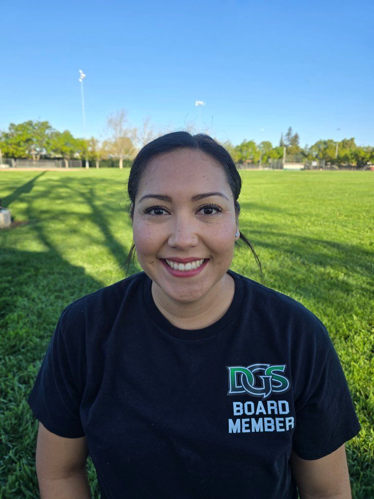 Football coach with arms crossed, standing on a football field. He wears a navy polo shirt and gray pants.