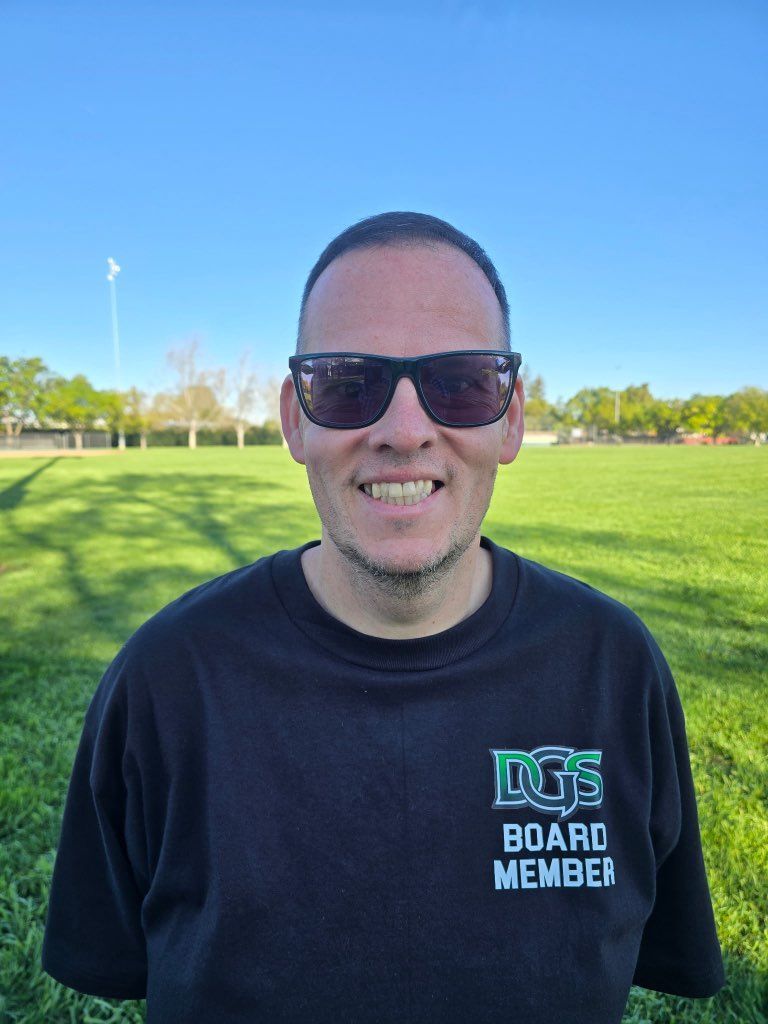 Man in green polo stands on a football field, arms crossed, stadium in background.