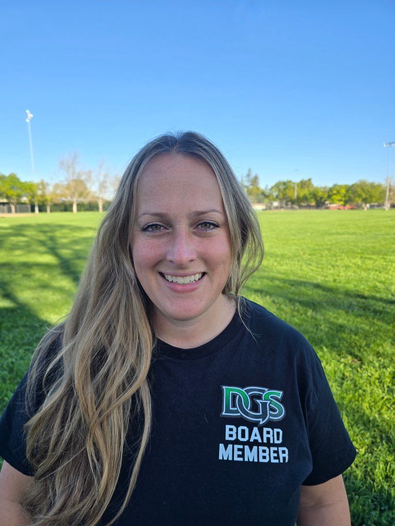 Football coach with arms crossed, looking stern on a sunny field, players in the background.